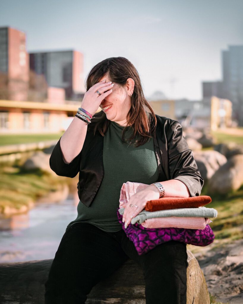 Linda moet om zichzelf lachen en zit op een bankje terwijl ze verschillende stoffen op schoot heeft liggen. Ze draagt haar eigen duurzame op maat gemaakte kleding.